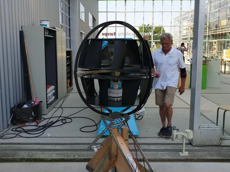 Een vroeg prototype van de turbine opgesteld op een houten frame bij een testlocatie, met een anemometer ernaast.