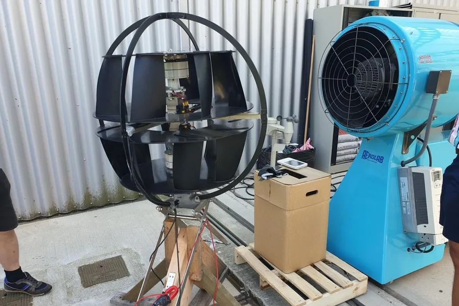 Een prototype wordt getest tegenover een grote windmachine, met meetapparatuur en een multimeter aangesloten.